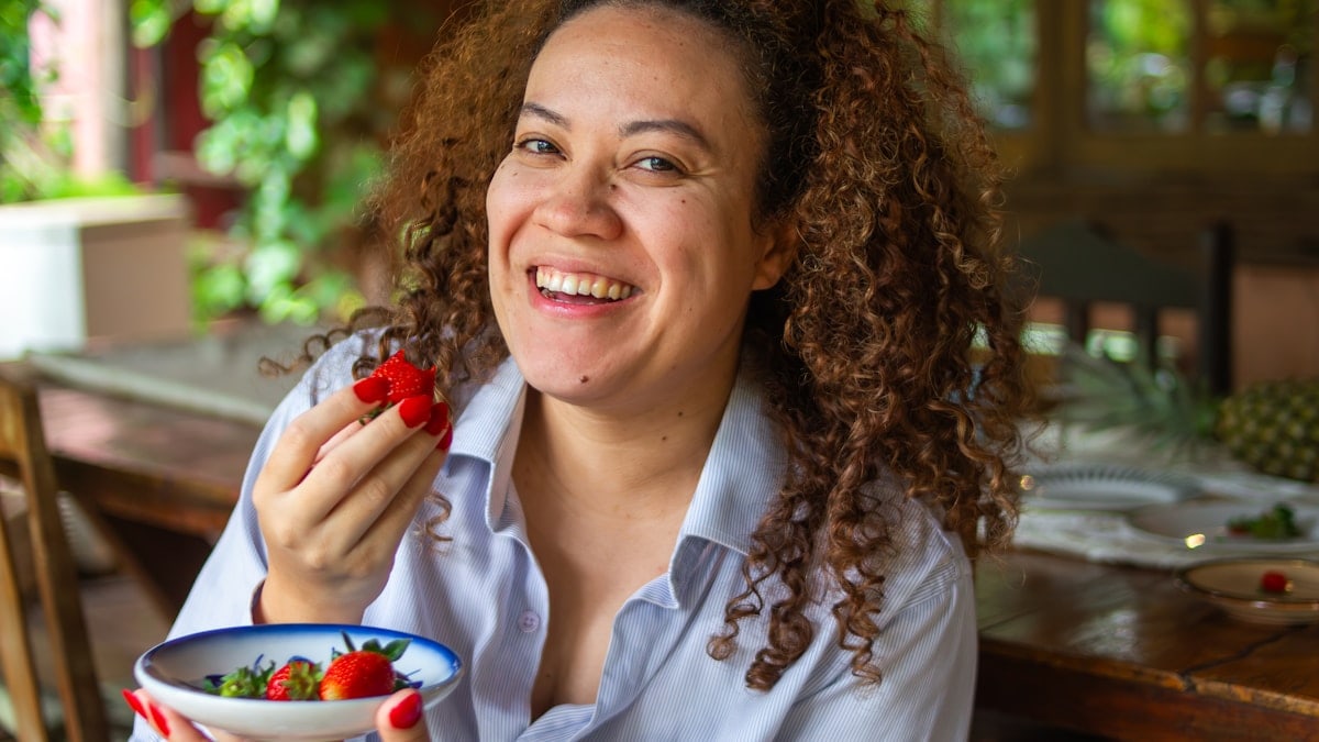 Smiling woman eating strawberries in a cozy restaurant with natural light and a warm lifestyle atmosphere.